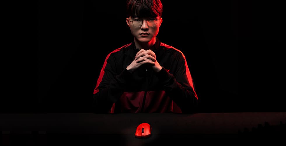 Joueur professionnel Faker portant des lunettes, éclairé en rouge et noir, les mains jointes devant une souris Razer DeathAdder V3 Pro