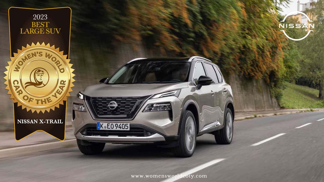 Nissan X-Trail gris clair roulant sur une route bordée de feuillage, avec médaille dorée du Women's World Car of the Year 2023 pour meilleur grand SUV