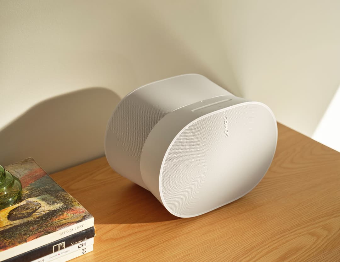 Enceinte intelligente Sonos de couleur blanche, design ovale arrondi, posée sur une table en bois à côté de livres