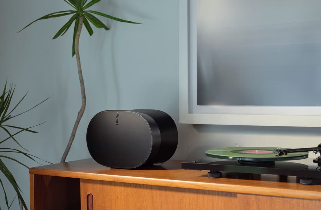 Enceinte Sonos Era 300 noire posée sur un meuble en bois à côté d'une platine vinyle, décor minimaliste avec plante verte et tableau