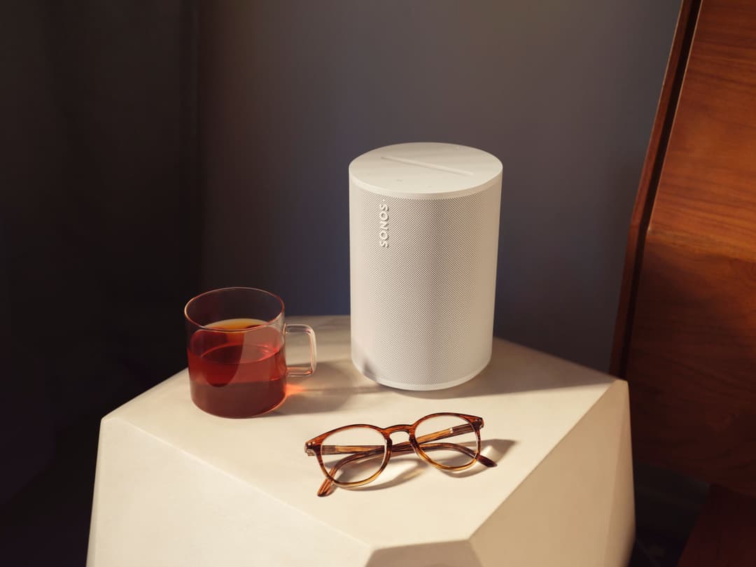 Enceinte Sonos Era 100 blanche posée sur un socle crème, à côté d'une tasse de thé et de lunettes de lecture