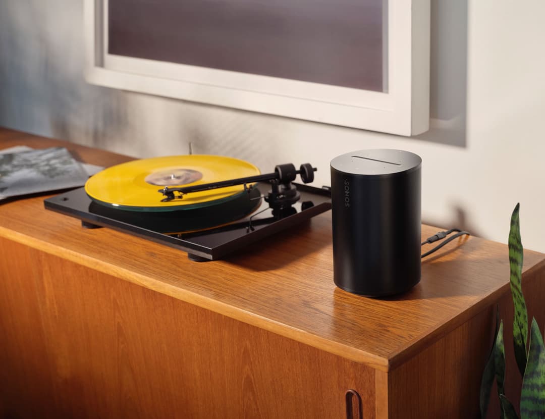 Sonos Era 100 noir cylindrique placé à côté d'une platine vinyle jaune sur un meuble en bois