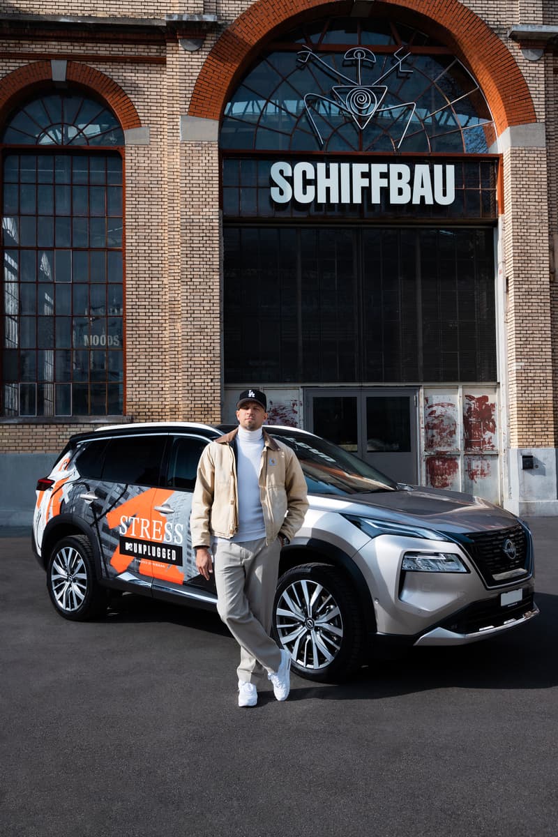 Le rappeur Stress posant devant un Nissan blanc et orange aux couleurs de son projet MTV Unplugged, devant un bâtiment en briques rouges