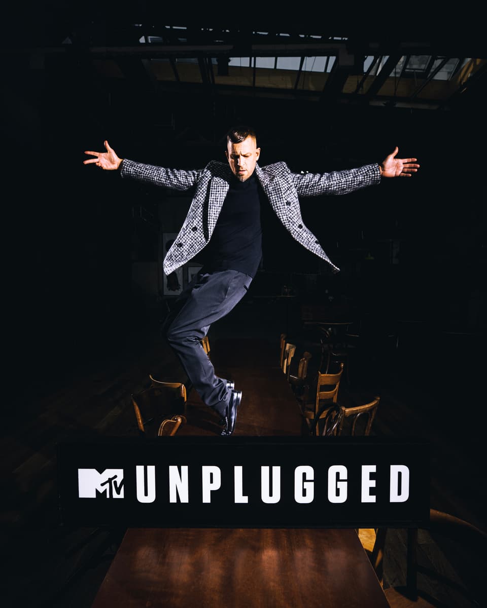 Le rappeur Stress saute les bras écartés sur scène, vêtu d'un blazer à carreaux blanc et noir, logo MTV Unplugged en bas de l'image