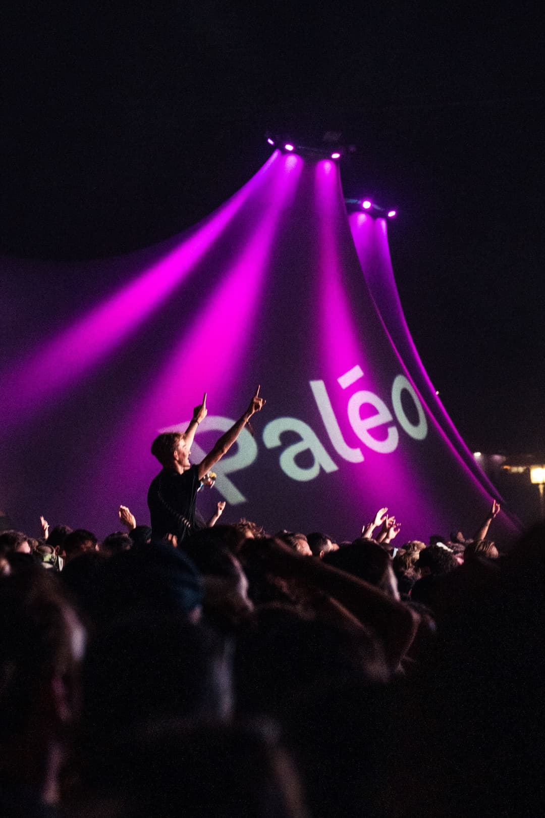 Artiste sur scène au Paléo Festival, bras levés face à la foule, éclairé par des projecteurs magenta