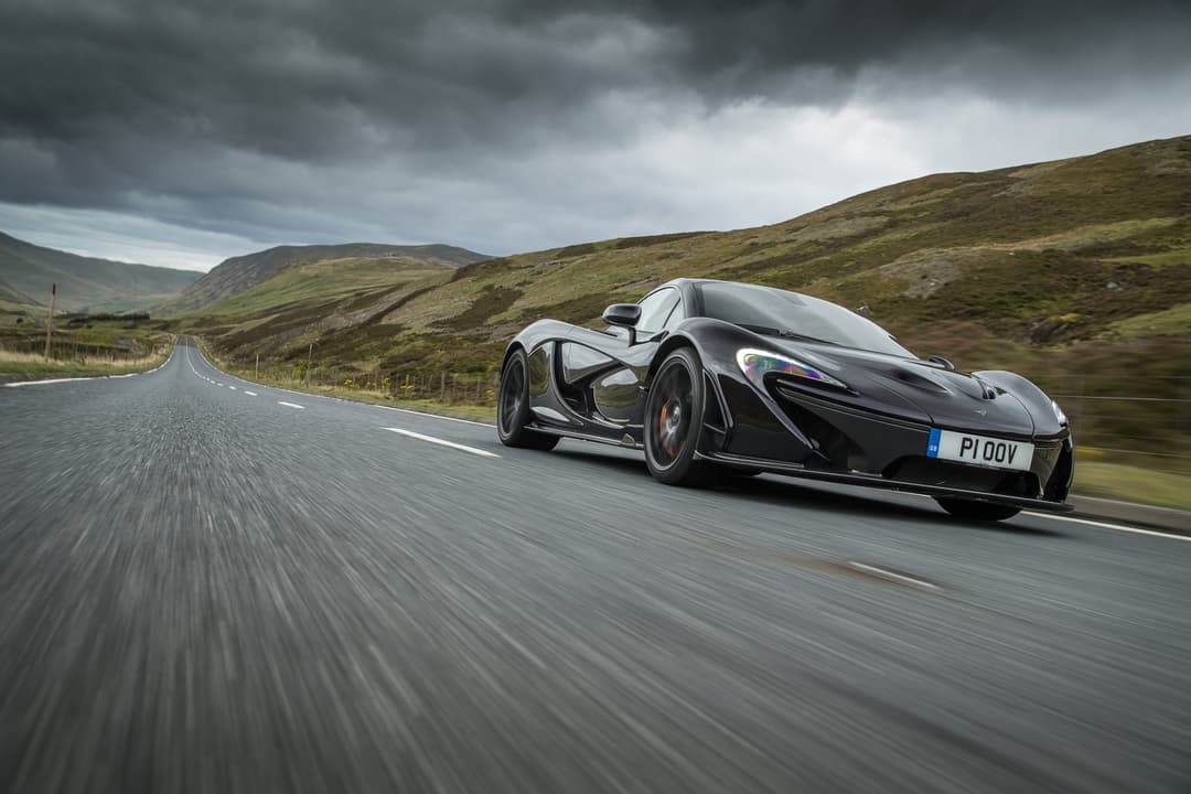 McLaren P1 noire en mouvement sur une route britannique, paysage montagneux verdoyant sous ciel nuageux
