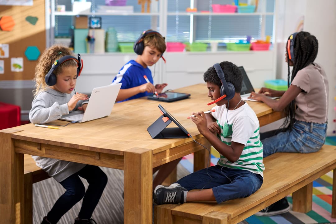 Quatre enfants portant des casques Logitech Zone Learn over-ear et on-ear travaillent sur des appareils numériques autour d'une table en bois dans une salle de classe