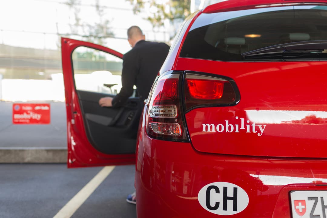 Voiture rouge Mobility avec logo visible, conducteur montant à bord, plaques d'immatriculation suisse