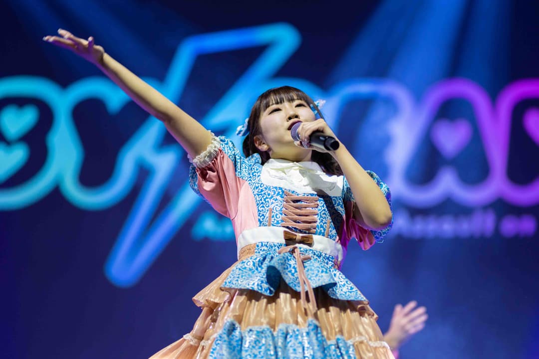 Jeune chanteuse en costume bleu et or chantant au microphone sur scène, bras levé, avec logo Polymanga flou en arrière-plan