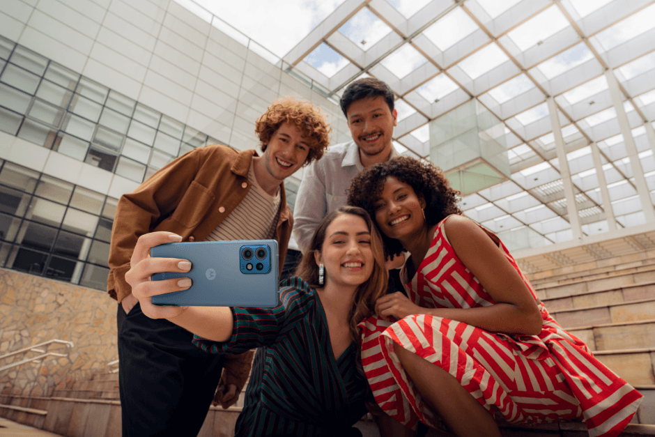 Quatre jeunes prennent un selfie avec le Motorola edge 40 pro bleu dans un bâtiment moderne aux plafonds vitrés