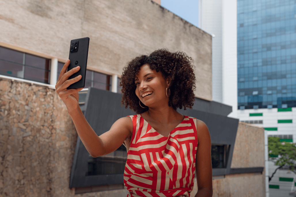 Femme tenant un Motorola Edge 40 Pro noir en main levée pour un selfie, vêtue d'un débardeur rouge à motifs