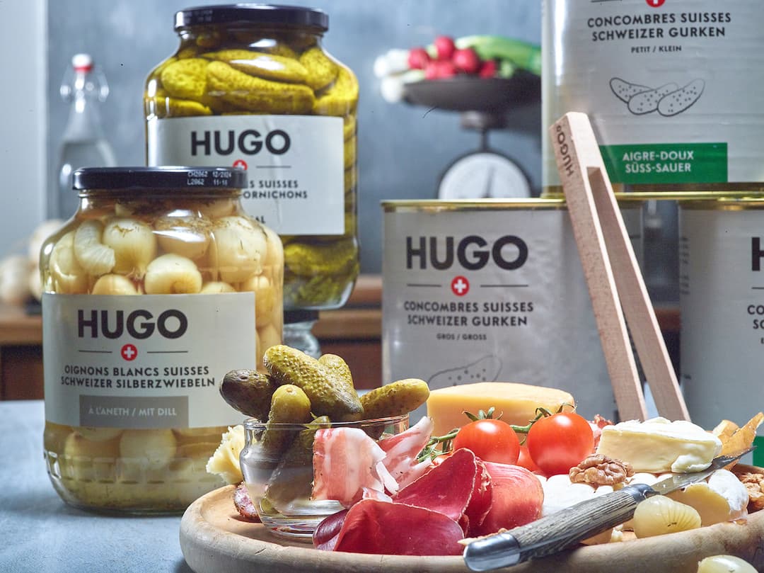 Produits HUGO suisses : bocaux de cornichons et oignons blancs, accompagnements et charcuterie locale sur planche de bois