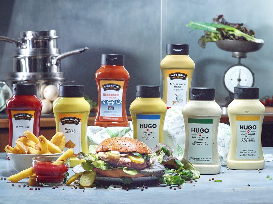Gamme de sauces HUGO Reitzel : bouteilles de ketchup, moutarde, sauce curry et tartare suisse, avec frites et burger en mise en scène