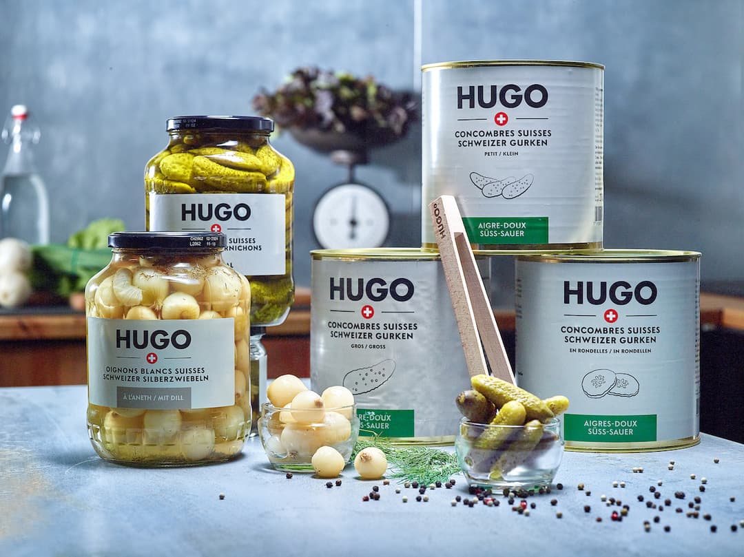 Gamme de produits HUGO : bocaux de cornichons et d'oignons blancs suisses, boîtes de concombre et cornichons aigre-doux, disposés sur un plan de travail