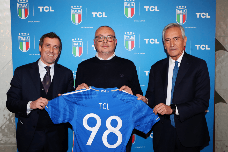 Trois hommes en costume tenant un maillot bleu de la Fédération italienne de football avec le logo TCL, devant un fond bleu avec logos Italia et TCL