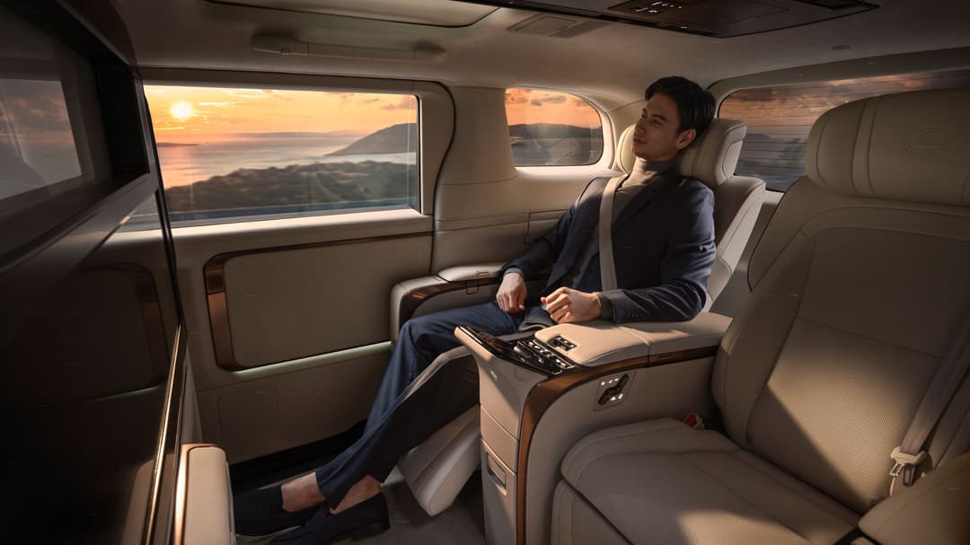 Intérieur spacieux du Lexus LM avec un passager assis dans des sièges beige, vue sur un paysage côtier au coucher de soleil par les vitres