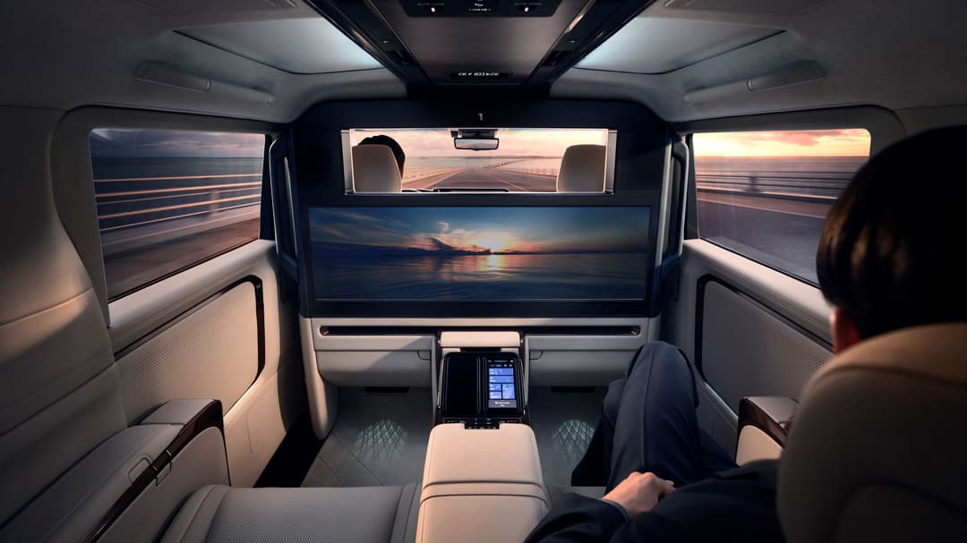 Habitacle du Lexus LM avec sièges blancs, grand écran panoramique HD affichant un coucher de soleil, et vue panoramique sur route au coucher du jour