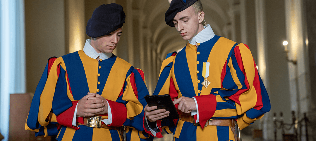 Deux gardes suisses pontificaux en uniforme traditionnel bleu, jaune et rouge consultent une tablette Samsung dans les couloirs du Vatican