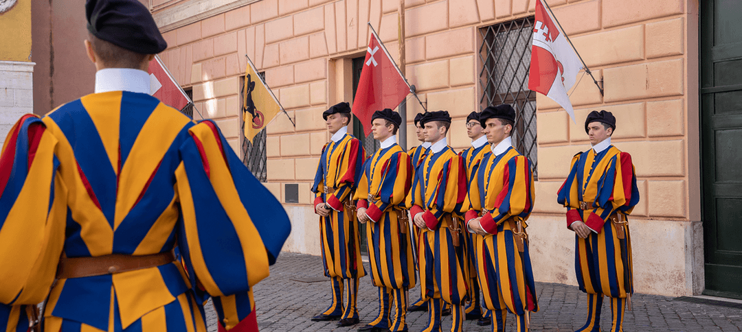 Gardes suisses pontificaux en uniforme bleu et jaune tenant des drapeaux devant une façade en pierre du Vatican
