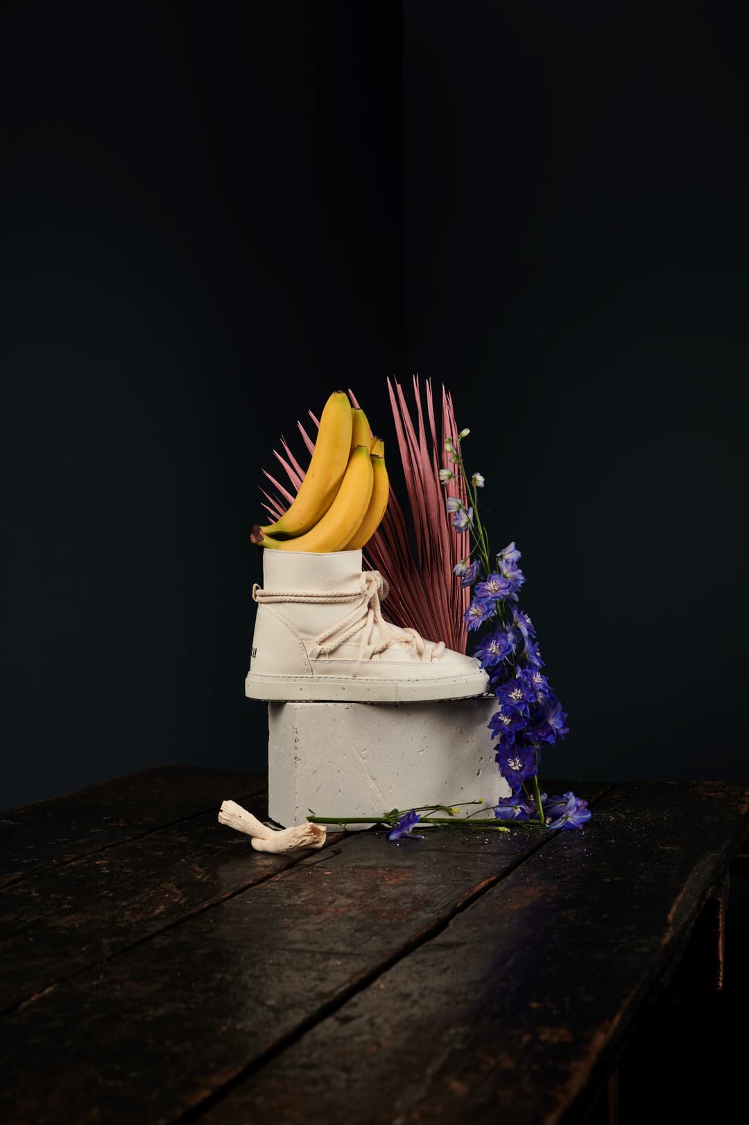 Chaussure INUIKII blanche transformée en composition artistique avec bananes jaunes, fleurs violettes et éléments naturels sur fond noir