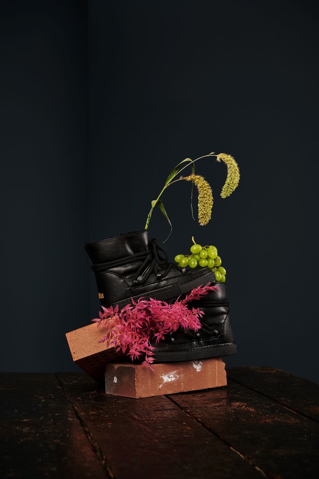 Botte noire INUIKII posée sur des briques, ornée de plants de bananier avec fleurs jaunes et grappes de raisin vert