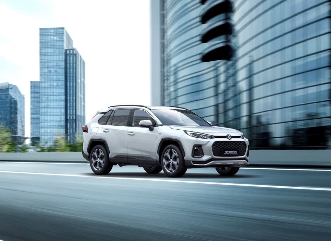 Suzuki Across blanc en profil, circulant devant des immeubles de verre modernes en milieu urbain