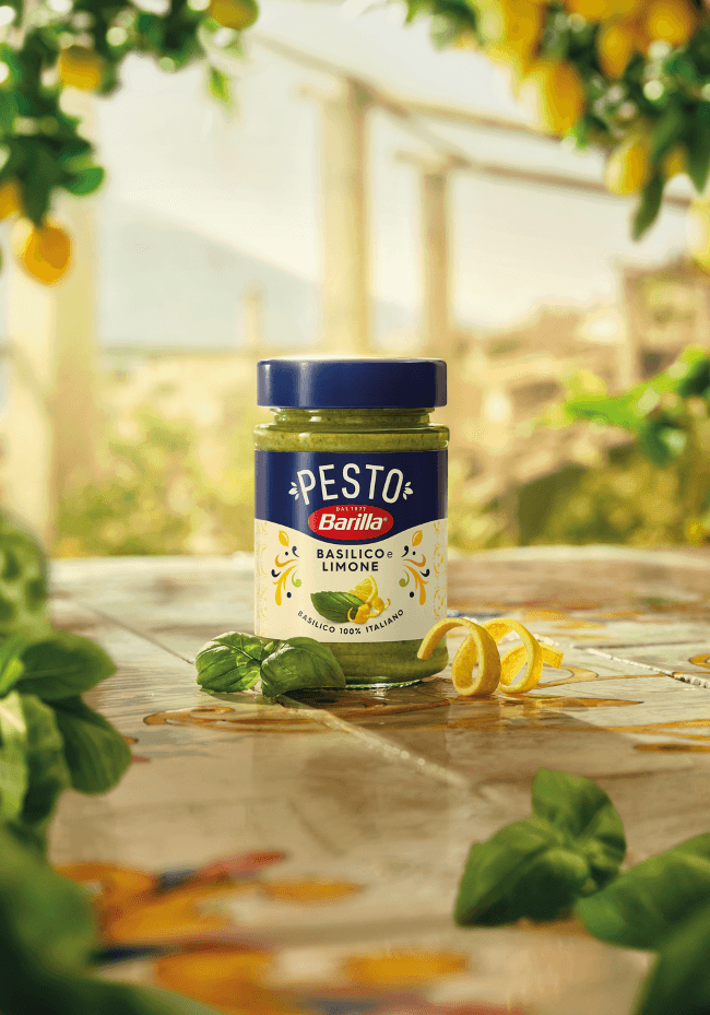 Pot de pesto Barilla Basilico e Limone entouré de feuilles de basilic frais et de zestes de citron sur un plan de travail ensoleillé