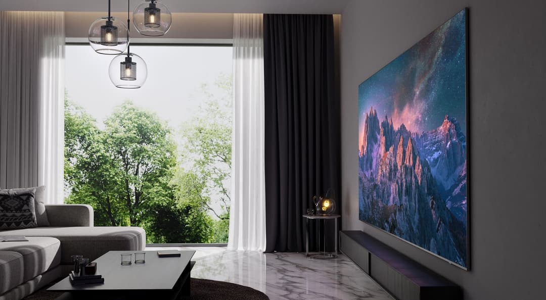 Téléviseur Samsung QLED 98 pouces affichant un paysage montagneux aux teintes roses et bleues, installé dans un salon moderne avec canapé gris et baie vitrée