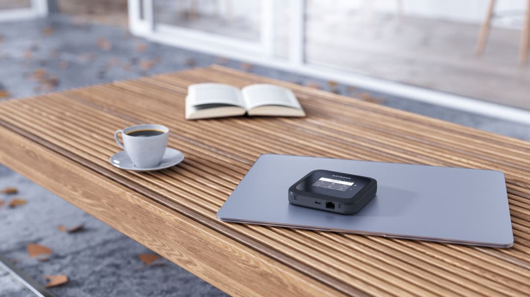 Routeur mobile NETGEAR M6 noir posé sur un ordinateur portable gris, sur une table en bois avec un café et un livre