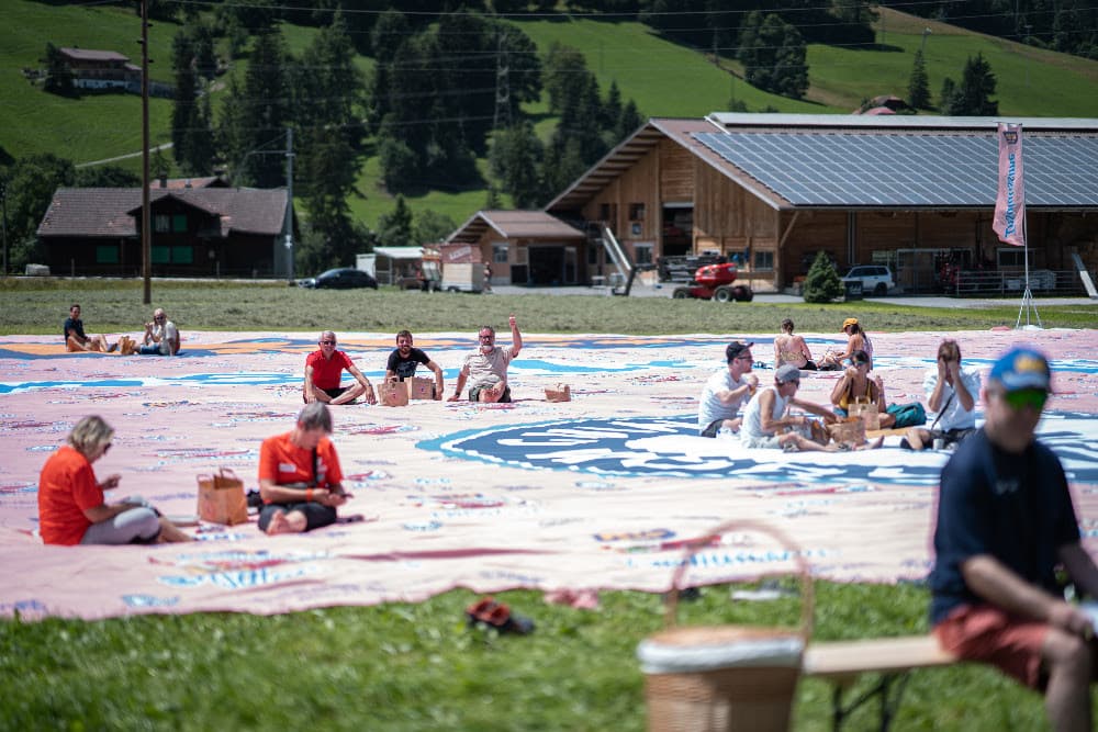 Grande nappe rose et bleu d'Insalatissime Rio Mare étendue sur un pré montagnard avec des participants assis dessus pour un pique-nique