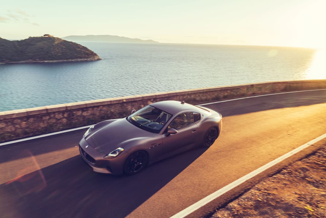 Maserati Gran Turismo Folgore grise circulant sur une route côtière au coucher du soleil, avec la mer et des îles en arrière-plan