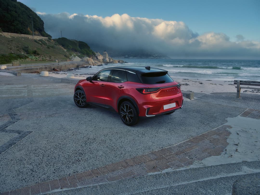 Lexus LBX rouge stationnée sur une plage, face à la mer et falaises sous ciel bleu nuageux