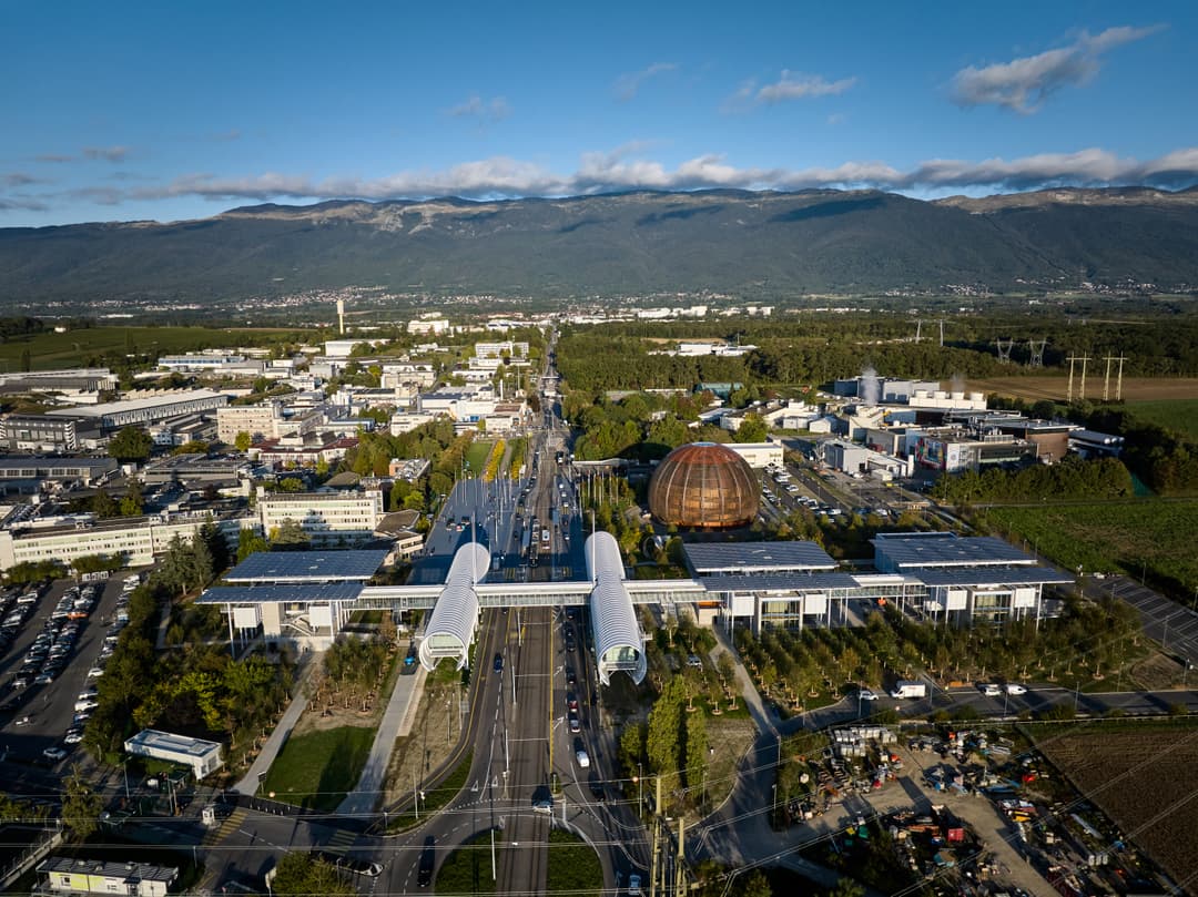 Aérienne du campus du Portail de la science du CERN, avec bâtiments modernes aux toits solaires, dôme métallique cuivré, arbres plantés et montagnes en arrière-plan