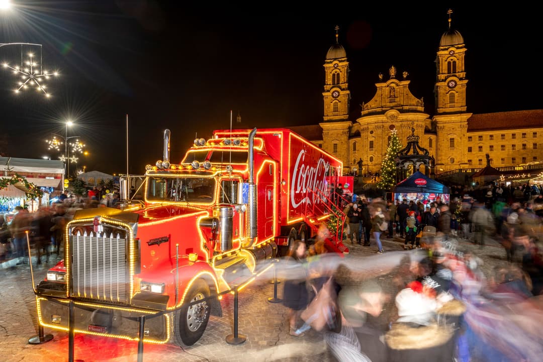 Le camion de Noël de Coca-Cola illuminé en rouge et or, stationné sur une place publique la nuit, avec des visiteurs autour et une église baroquée éclairée en arrière-plan