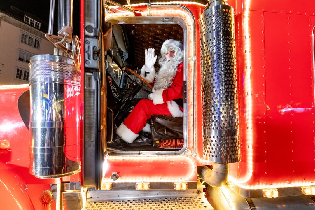 Père Noël en costume rouge assis dans la cabine du camion de Noël Coca-Cola illuminé, saluant par la fenêtre