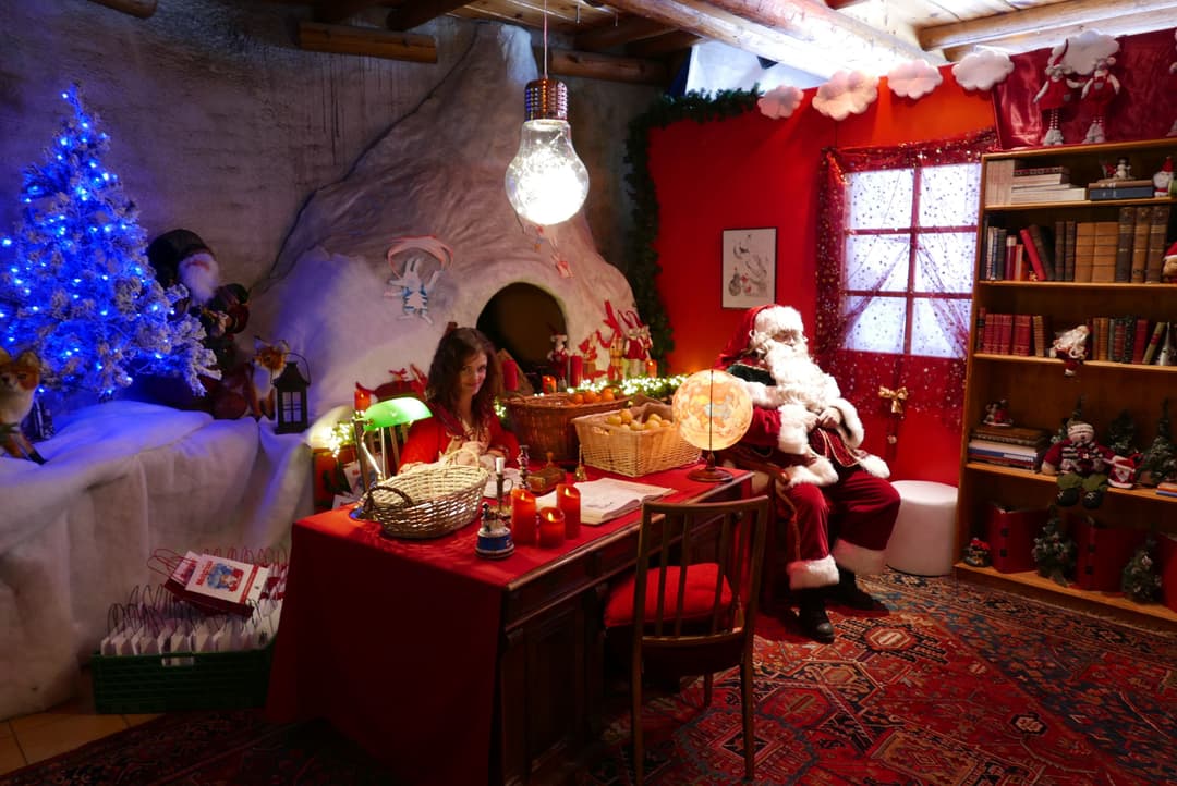Intérieur de la Maison du Père Noël illuminé en rouge et bleu, avec sapin luminescent, père Noël à son bureau et enfant visiteur