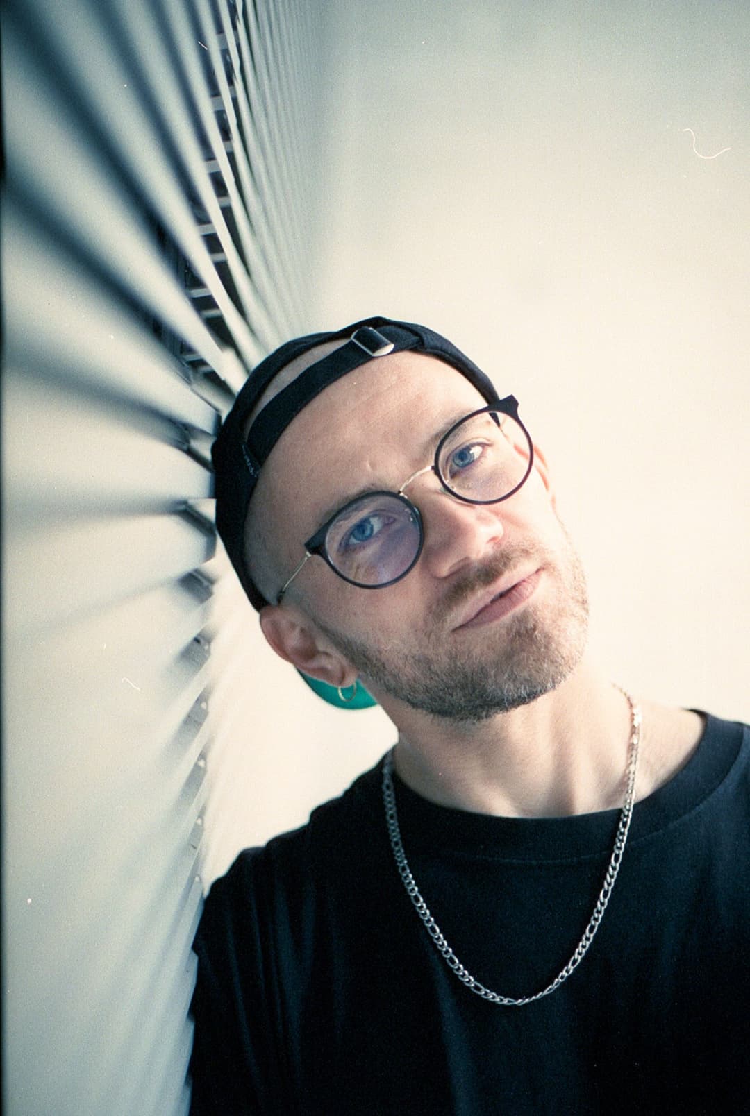 Portrait de Rylsee portant des lunettes rondes, une casquette noire et un collier chaîne, appuyé contre une façade blanche