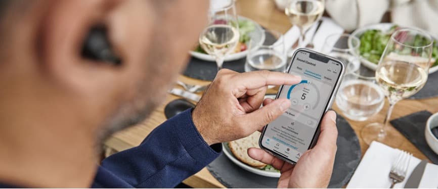 Homme tenant un smartphone affichant l'application de contrôle d'un casque audio, assis à table avec verres et assiettes