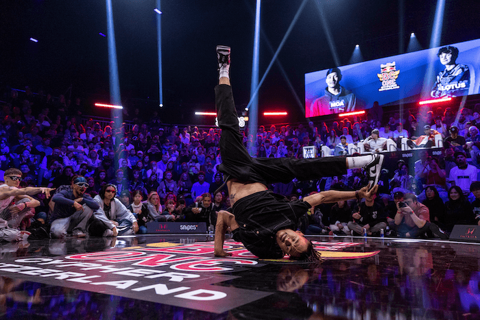 Danseur de breaking en figure acrobatique sur la piste du Red Bull BC One City Cypher, devant une foule éclairée par projecteurs bleus et rouges