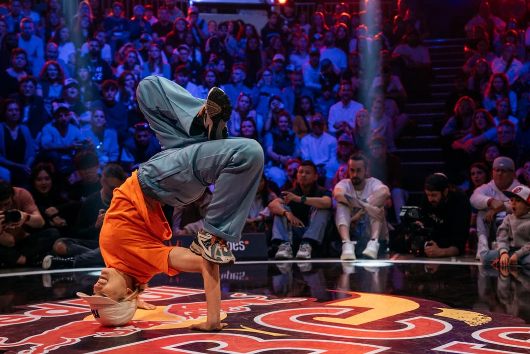 Danseur de breaking en costume orange effectuant un freeze sur la tête, entouré d'une foule de spectateurs sous éclairage bleu et rouge