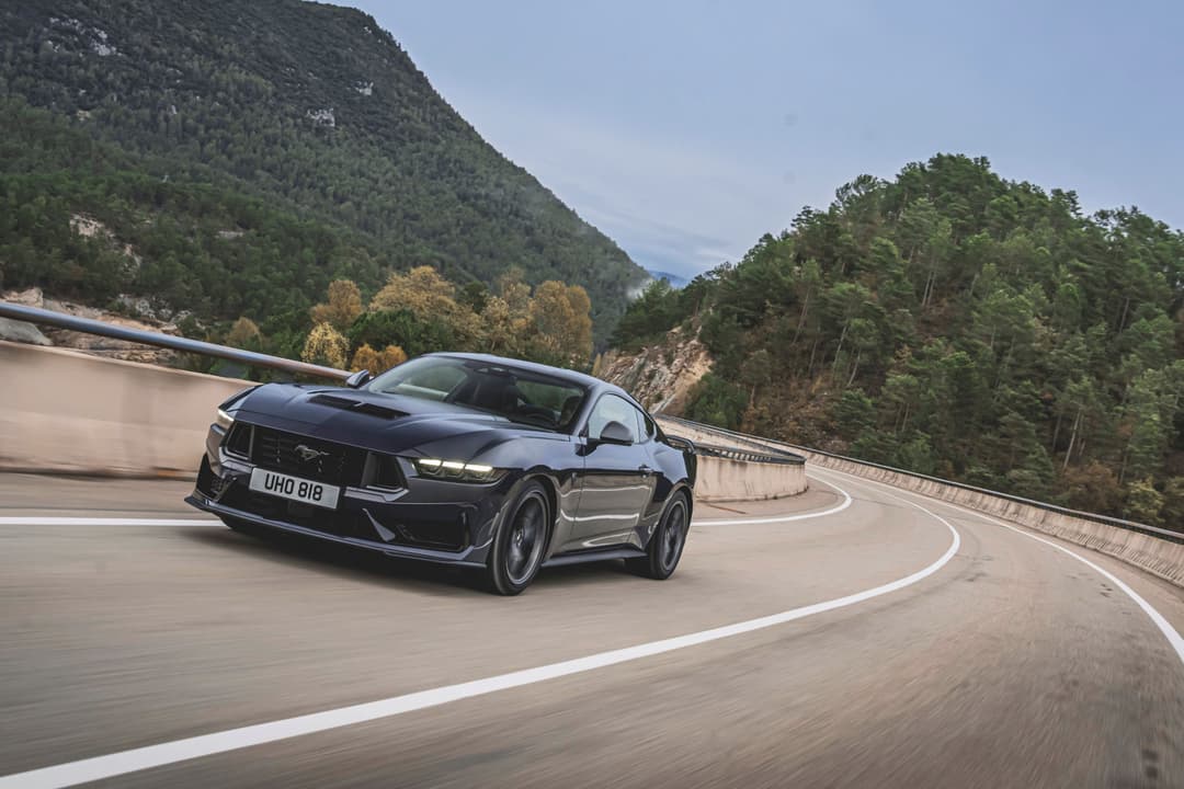 Ford Mustang noire en virage sur une route de montagne sinueuse, entourée de forêts et de falaises