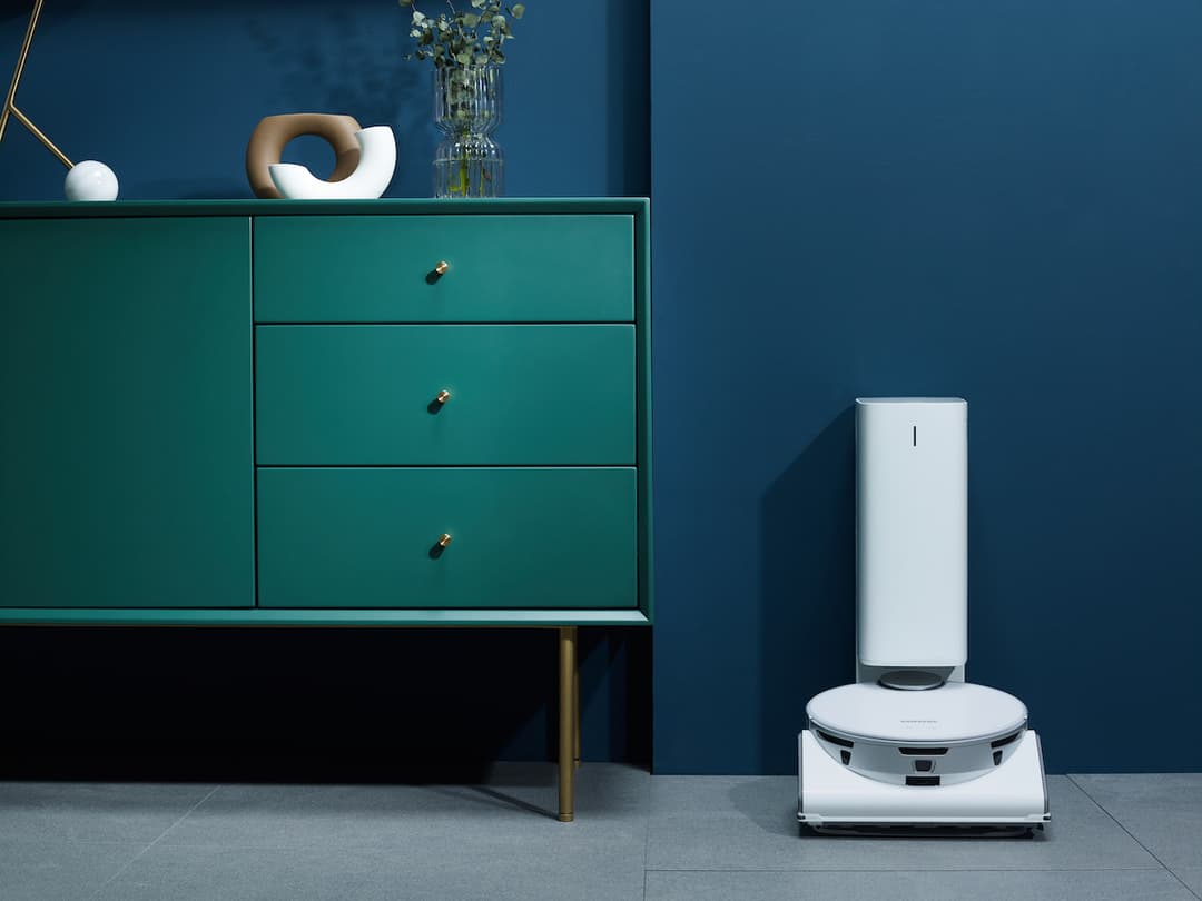 Robot aspirateur blanc Samsung avec station de vidage automatique dans un intérieur moderne aux murs bleu foncé et meuble turquoise