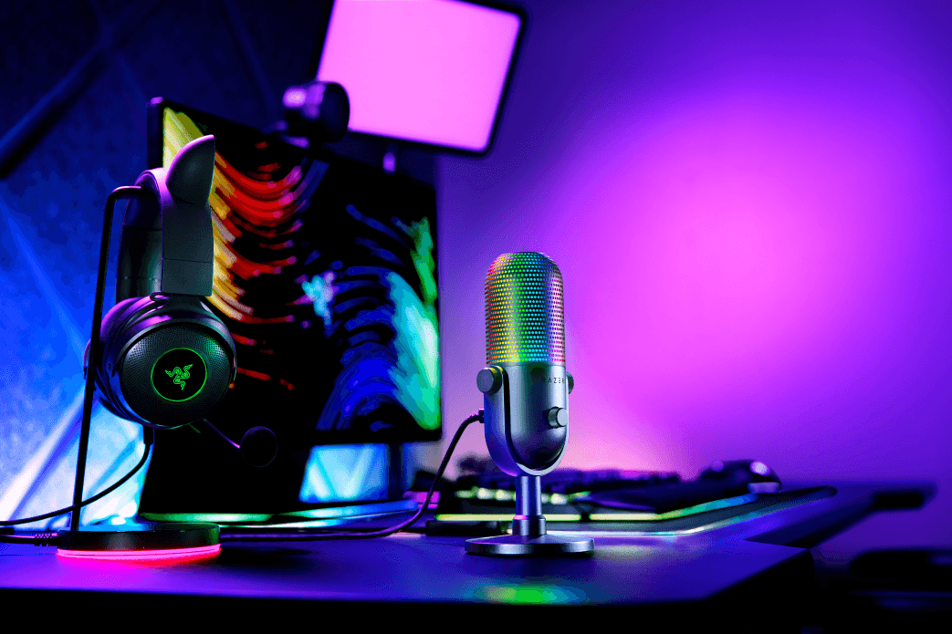 Microphone Razer Seiren V3 Chroma avec grille illuminée en dégradé de couleurs, casque gaming Razer et écran en arrière-plan, ambiance RGB violette et bleue