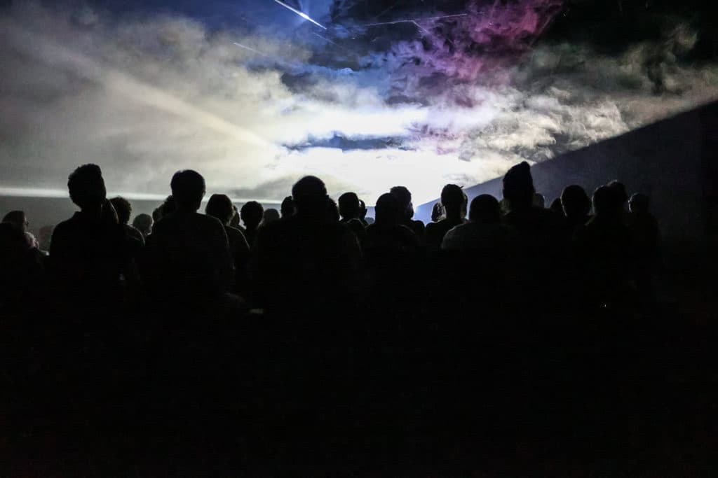 Silhouettes d'une foule face à un ciel dramatique aux teintes bleues et violettes, illuminé par des effets lumineux
