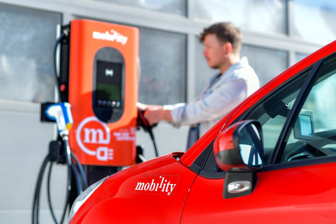 Jeune homme rechargeant une voiture électrique rouge Mobility sur une borne de car sharing orange