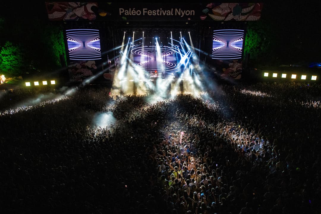 Scène principale du Paléo Festival Nyon de nuit, avec foule dense, projecteurs bleus et blancs, écrans géants aux motifs hypnotiques, verdure éclairée en arrière-plan
