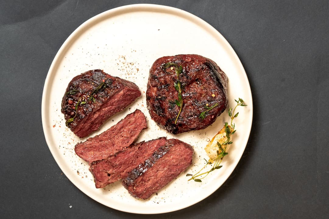 Planted.steak à base de plantes, tranché et entier, cuit et croûté, garni d'herbes aromatiques sur assiette blanche