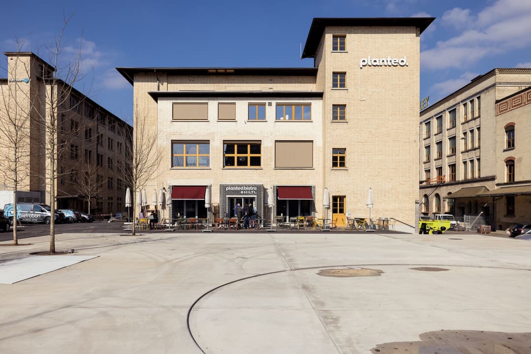 Bâtiment beige de Planted à Kemptthal avec logo blanc, terrasse en rez-de-chaussée, immeubles d'habitation alentour, place publique dégagée