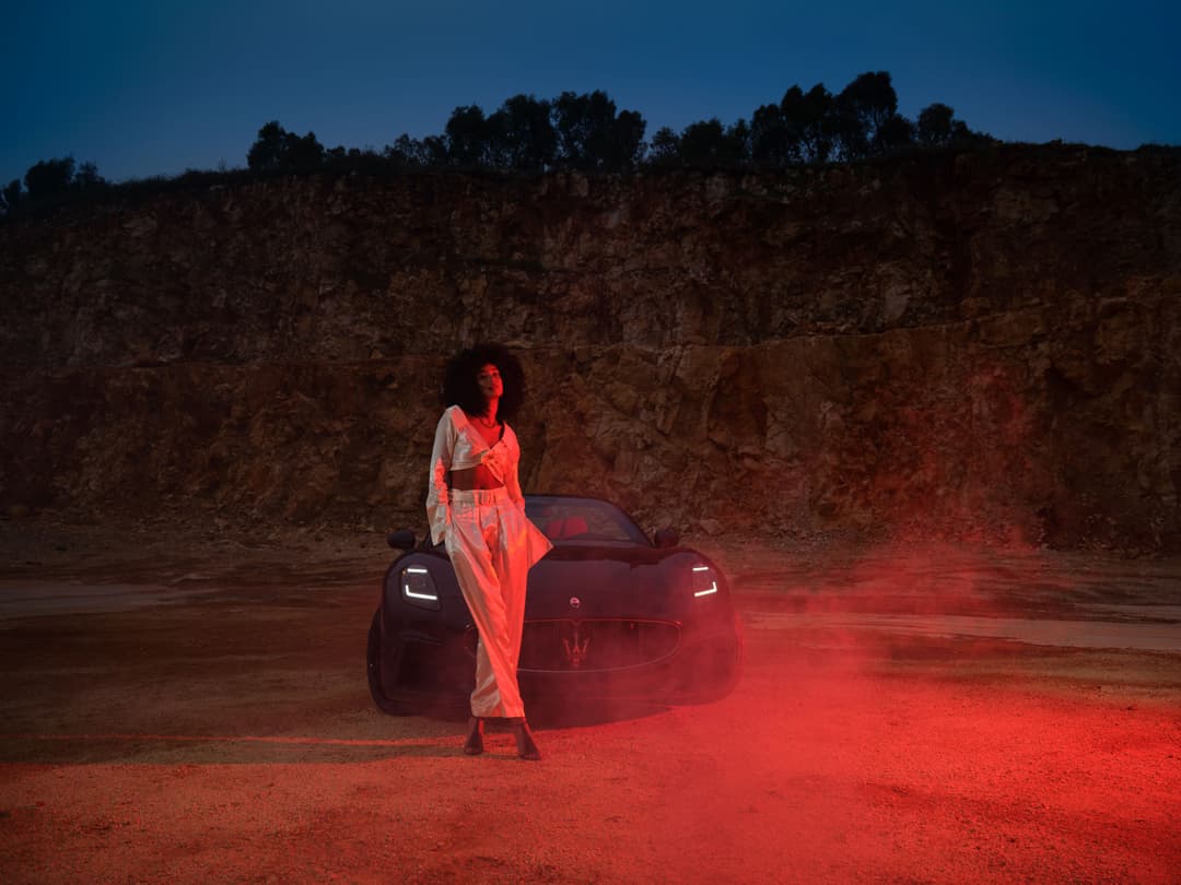 Maserati GranCabrio décapotable rouge illuminée la nuit, avec une femme en robe orange posée devant, dans un paysage rocheux