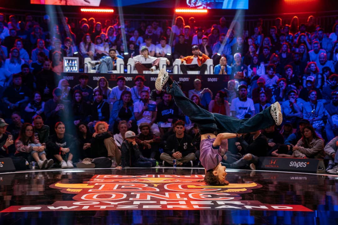 Danseur de breaking en figure acrobatique sur la piste du Red Bull BC One, entouré de spectateurs et du jury